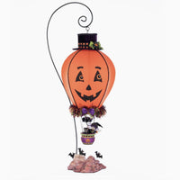 Full-length image of the Katherines Collection Halloween Ghoulish Hot Air Balloon with glowing light, pumpkin face wearing a top hat with plaid fabric trim, witch and cat in basket and bats flying at the base