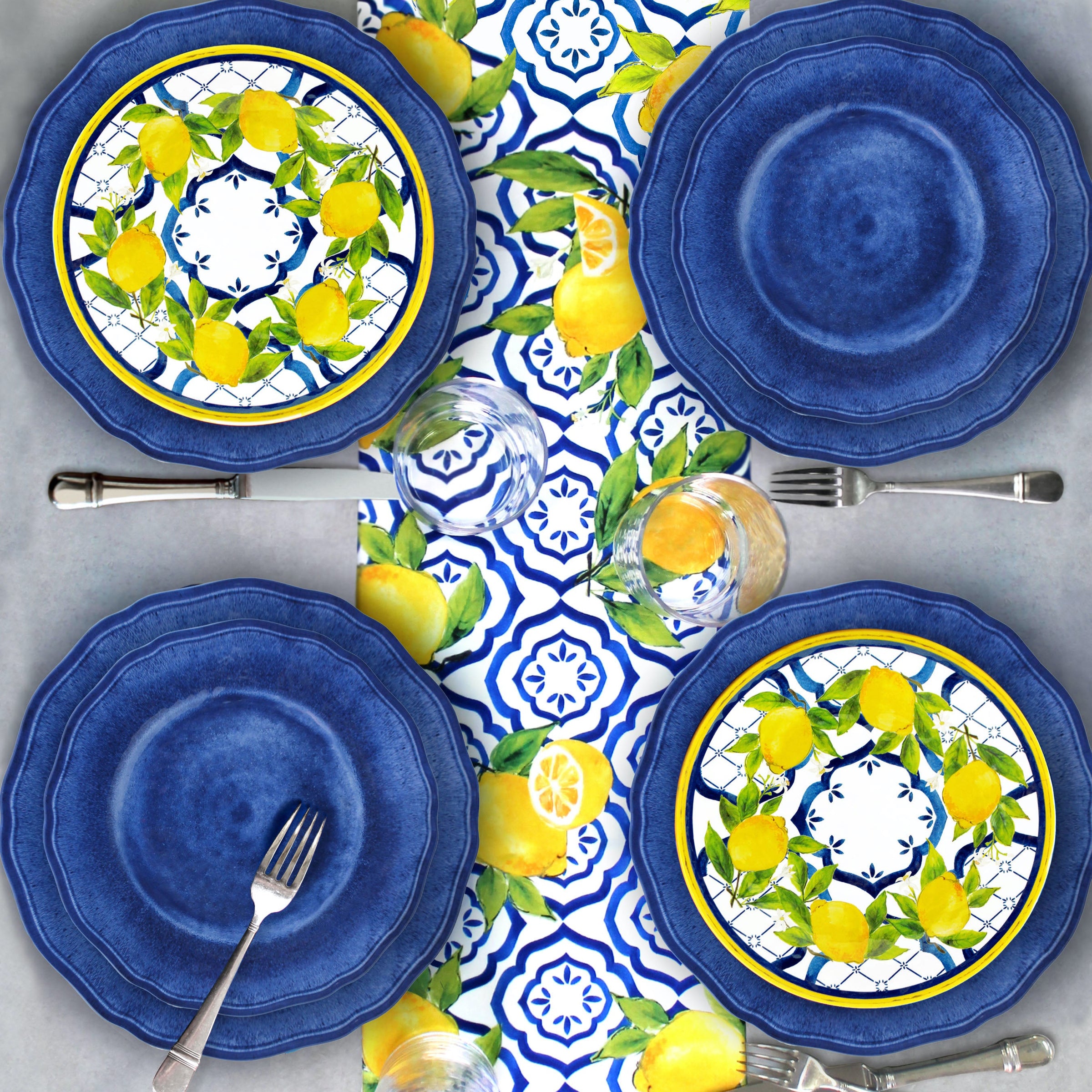 Set of le cadeaux Campania Blue blue dinner and salad plates with le Cadeaux Palermo lemon design salad plates on a matching Le Cadeaux Palermo tablecloth, Santorini Clear tumblers arranged on a gray surface.