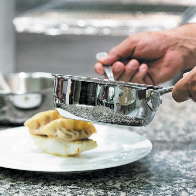 Chef holding a Cristel stainless steel pot over a plate with food on a kitchen counter.