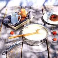 Stainless steel pan with wooden spoon on a wooden surface with fruits and a cake.