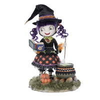 Front-facing view of Katherine's Collection Poppy Broomson Witch standing over her cauldron reading a spell book, with pumpkins at her feet in an orange and black buffalo plaid dress and jeweled accents. and black witches hat