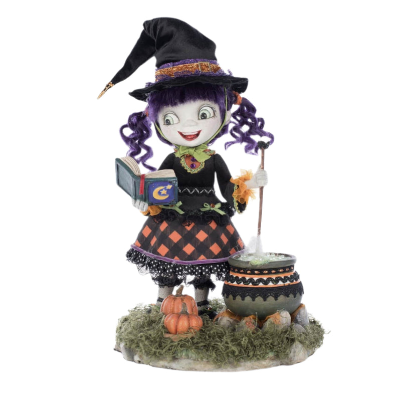 Front-facing view of Katherine's Collection Poppy Broomson Witch standing over her cauldron reading a spell book, with pumpkins at her feet in an orange and black buffalo plaid dress and jeweled accents. and black witches hat