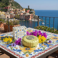 Decorative table with Rudy Profumi Riviera Mediterranean inspired bath soap, and flowers on a scenic background of a coastal town.