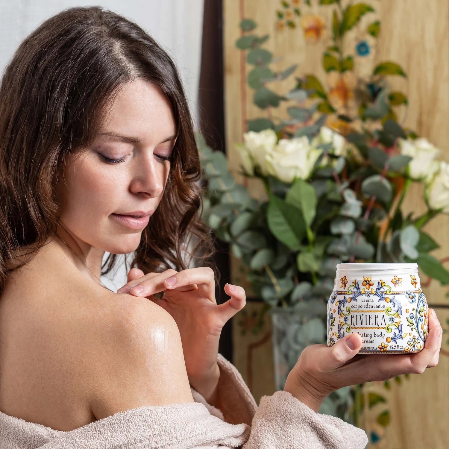 Woman applying Floral oriental Italian body moisturizer cream to her shoulder with a decorative jar in a home setting