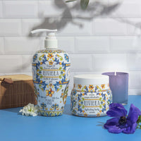 Riviera skincare products including a bottle, jar, and candle on a blue surface with flowers.