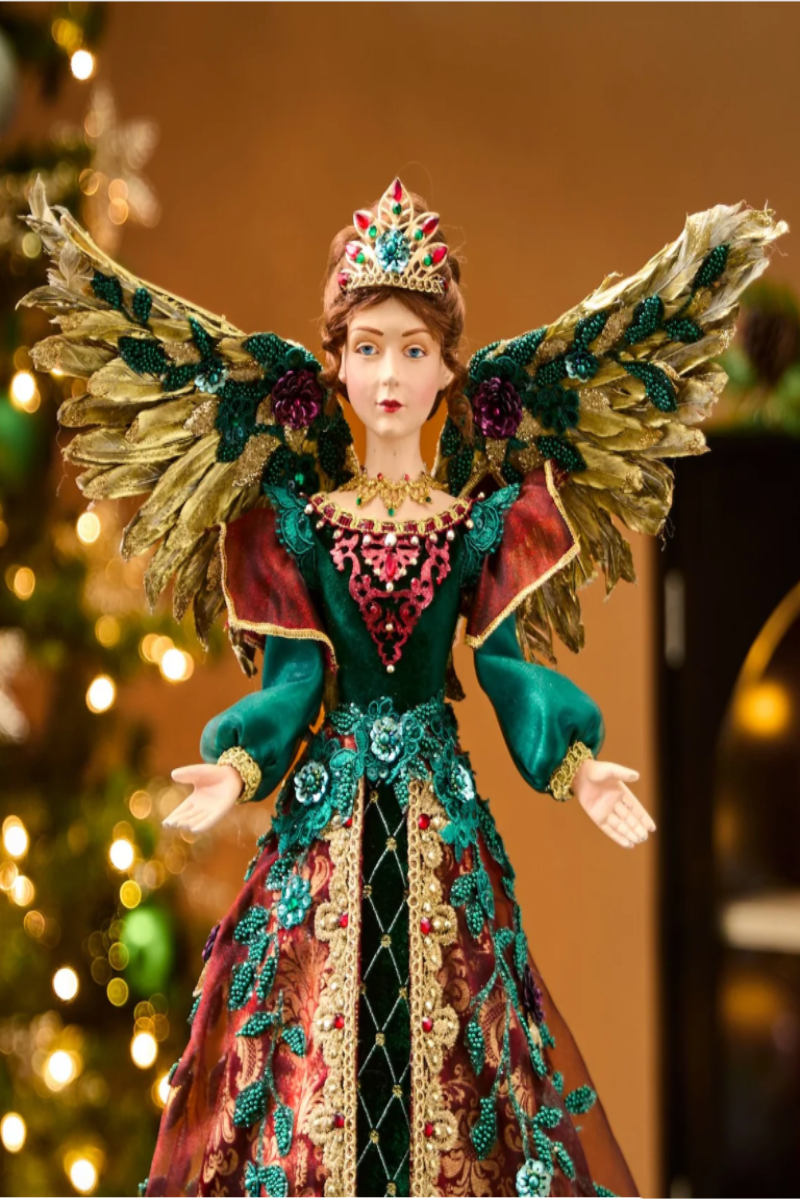 An ornate Christmas angel doll with large feathered wings, a jeweled crown, and an elaborate teal and burgundy gown stands in front of a softly blurred Christmas tree and lit holiday decor.